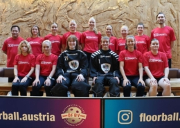 Das österreichische Damen-Nationalteam in roten Trikots, die beiden Torhüterinnen in Schwarz, aufgestellt in der Halle vor den Bannern des Verbands.